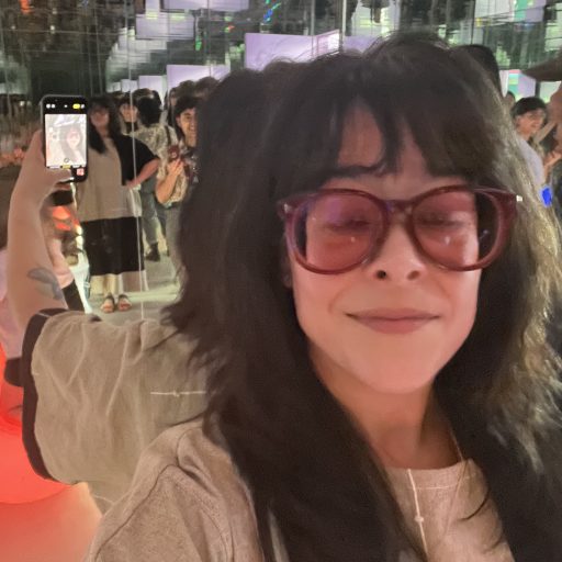 Light skinned brunette woman wearing pink framed glasses with pink lenses. She is standing in a room full of mirrors. Her eyes are closed because she blinked while taking the picture.